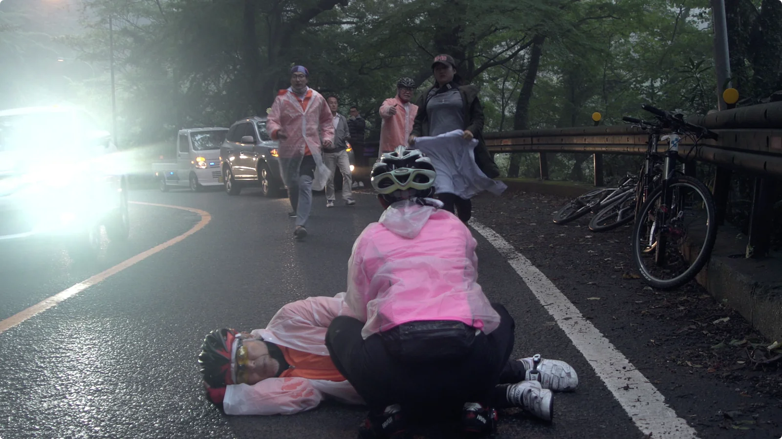 도로가에 멈춘 라이더들과 자전거들. 우비를 입은 한 사람 곁에 동료들이 모여 있다.